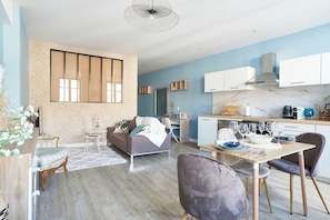 Een moderne woonkamer met een bank, een eettafel met stoelen, een keuken met kasten en een fornuis, en een groot raam met jaloezieën.
