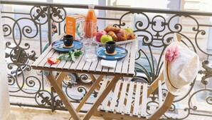 Un balcón con una mesa dispuesta para el desayuno, que incluye croissants, manzanas y jugo, junto con un sombrero decorativo y una rosa.