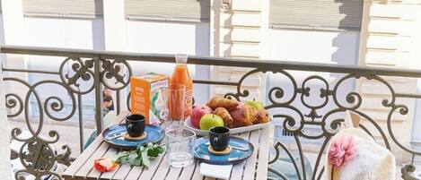 A balcony with a table set for breakfast, including croissants, apples, and juice, with a decorative hat and a rose.