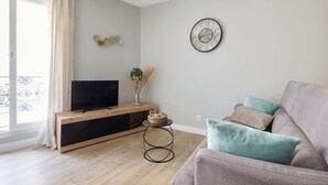 Una sala de estar con un televisor sobre una consola de madera, un reloj de pared redondo y un sofá gris con cojines.