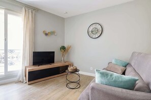 Ein Wohnzimmer mit Fernseher auf einer Holzkonsole, einer runden Wanduhr und einem grauen Sofa mit Kissen.