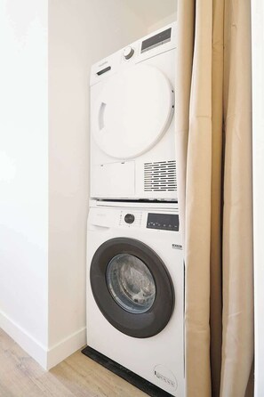 Une machine à laver compacte, de couleur blanche et à chargement frontal, placée dans un coin d’une pièce dont le sol est en bois et qui est dotée d’un rideau beige.