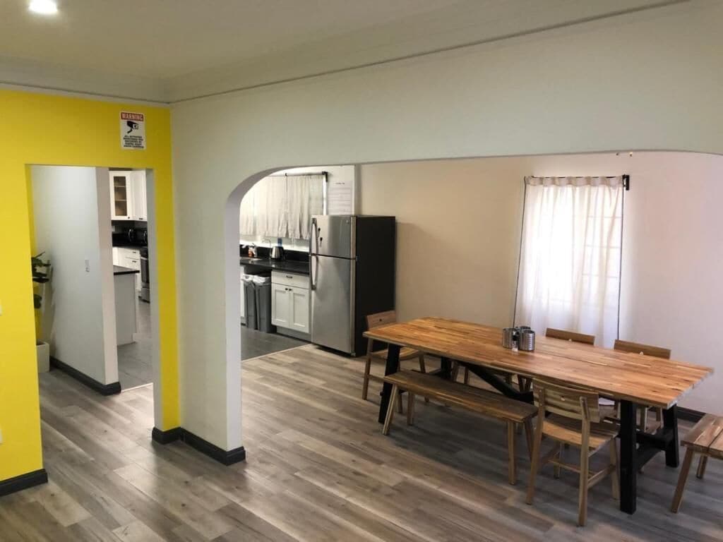 A dining area with a wooden table and benches, a kitchen in the background, and a yellow wall with a warning sign.