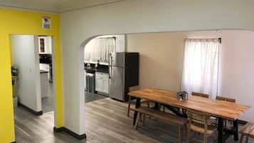 A dining area with a wooden table and benches, a kitchen in the background, and a yellow wall with a warning sign.