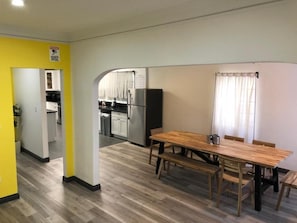 A dining area with a wooden table and benches, a kitchen in the background, and a yellow wall with a warning sign.