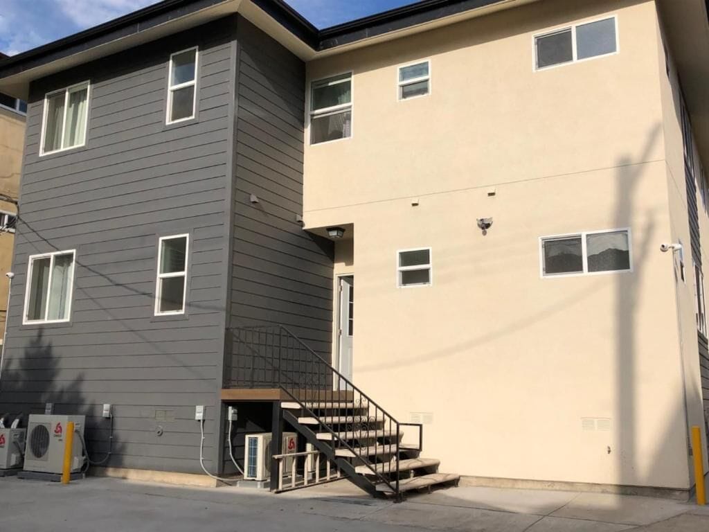 A two-story building with a grey and beige exterior, a visible staircase, and outdoor air conditioning units.
