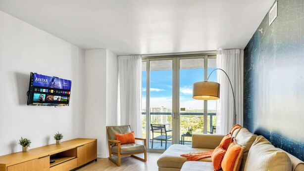 A modern hotel room with a large window, a flat-screen TV, a wooden desk, a sofa, and a chair. - Spacious 1BR - Bay View (Hallandale Beach)