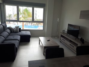 A modern living room with a sofa, ottoman, coffee table, TV stand, and a view of a swimming pool and buildings. - Apartment Mirador de Villamartin (Orihuela)