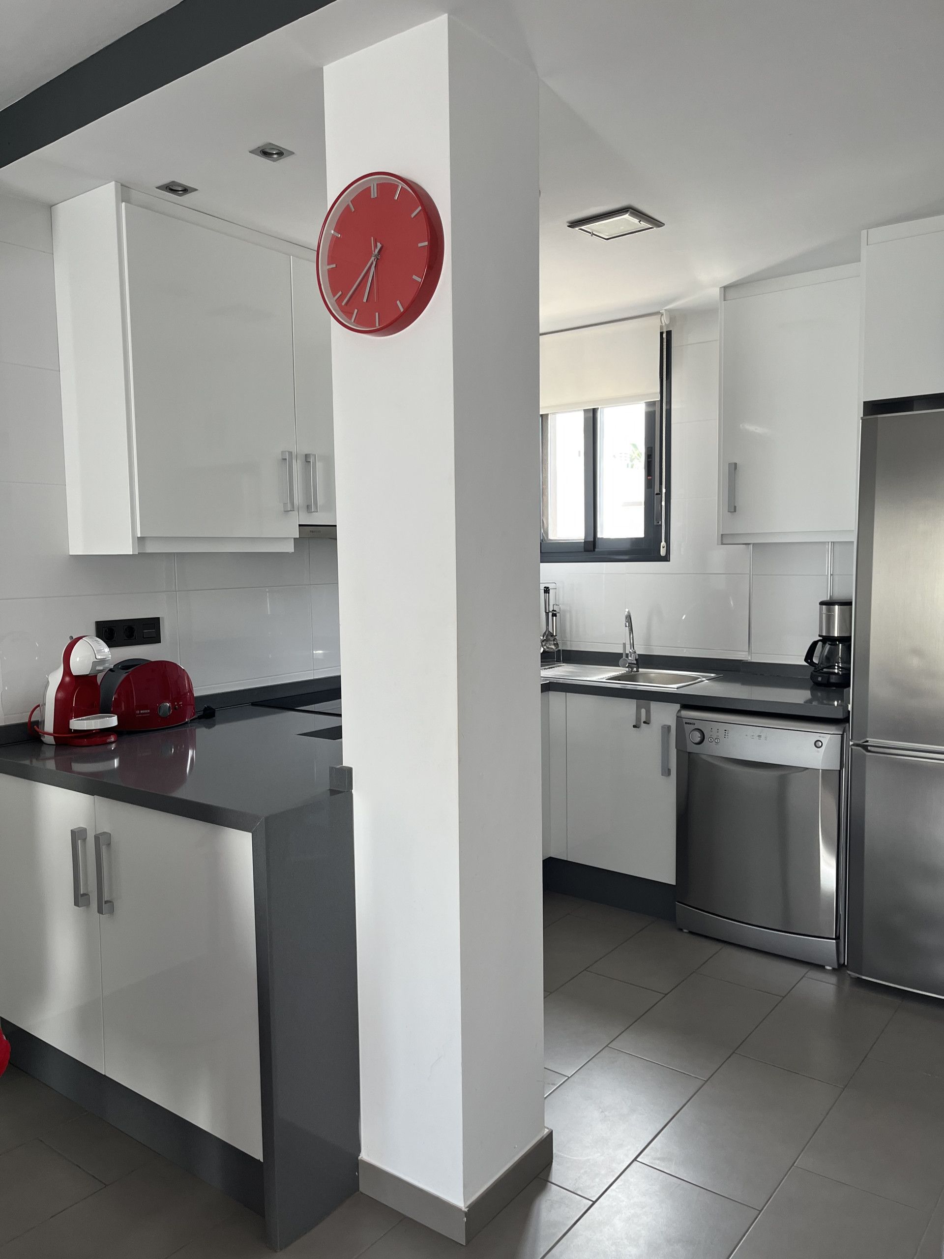 Una cocina moderna con armarios blancos, un refrigerador de acero inoxidable, un lavavajillas y un reloj rojo en la pared.