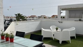 A rooftop terrace with a glass table and chairs, a small garden area with potted plants, and a view of the cityscape.