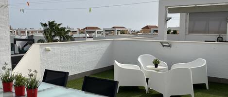 A rooftop terrace with a glass table and chairs, a small garden area with potted plants, and a view of the cityscape.