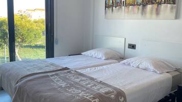 A double bed with a beige blanket, a cityscape painting on the wall, and a large window with a view of greenery and buildings.