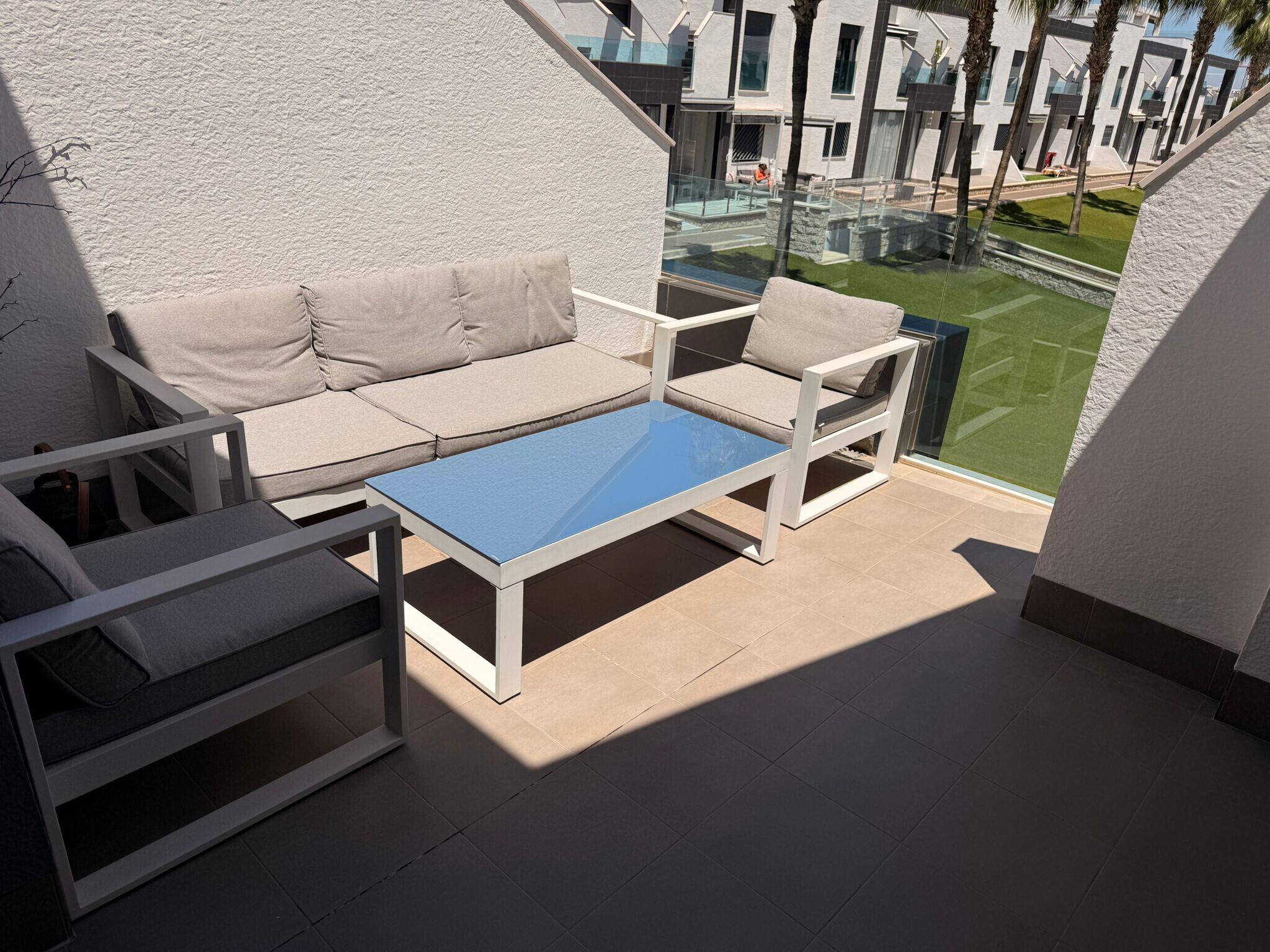 A balcony with a sofa and chairs, a glass table, and a view of a modern building and greenery.