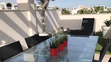 Una mesa con cubierta de vidrio y sillas negras, rodeada de macetas y un árbol.