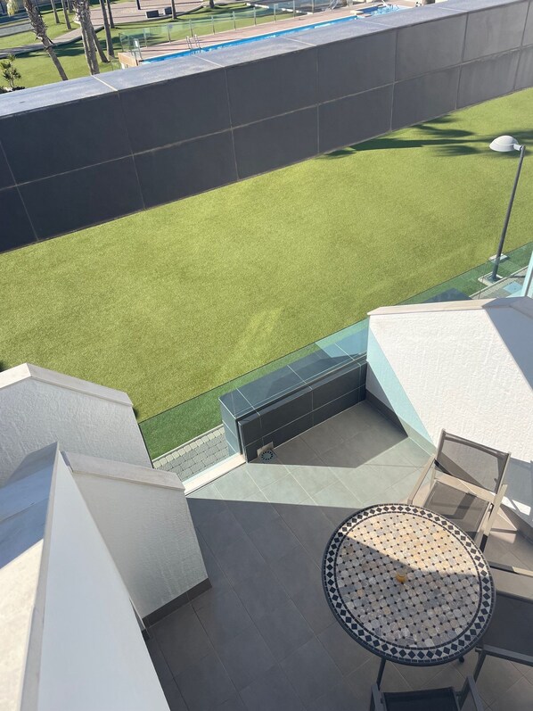 A rooftop terrace with a view of a green lawn, a tiled floor, and a round table with a patterned top.