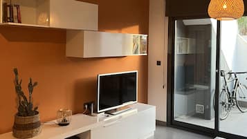 Un salon moderne avec un téléviseur à écran plat posé sur une console blanche, une porte vitrée donnant sur un balcon et un luminaire suspendu.