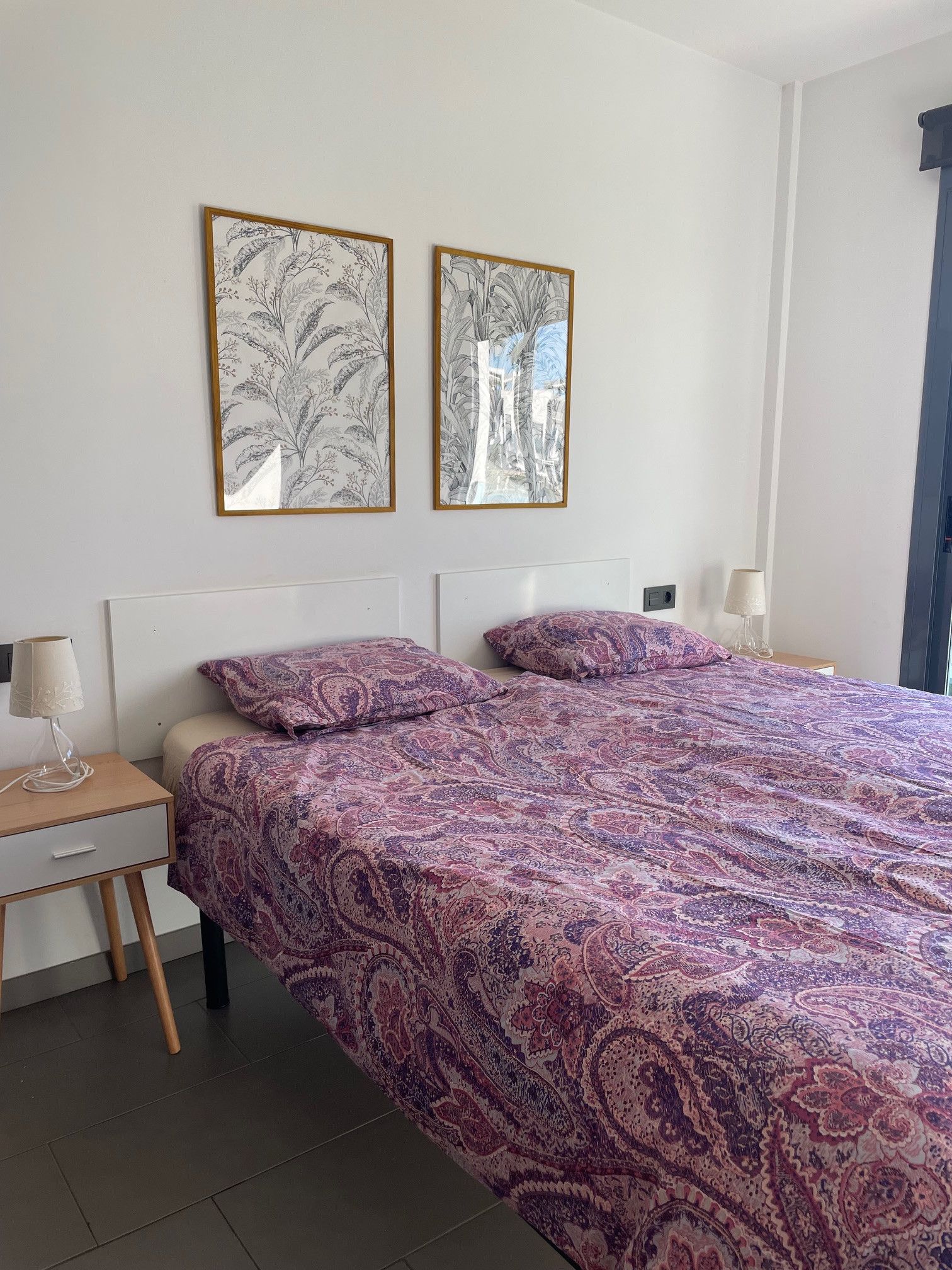 Une chambre à coucher avec un lit, deux reproductions botaniques encadrées accrochées au mur, une table de chevet avec une lampe et une commode.