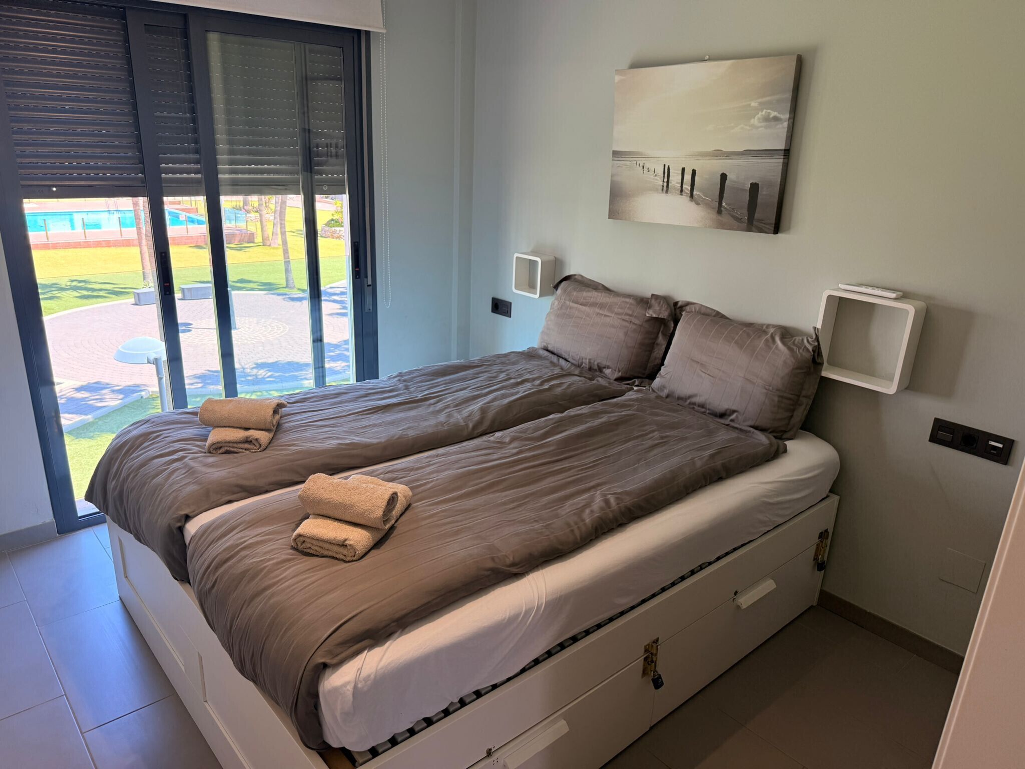Una cama doble con vistas a una piscina y un jardín, a través de un ventanal corredero.