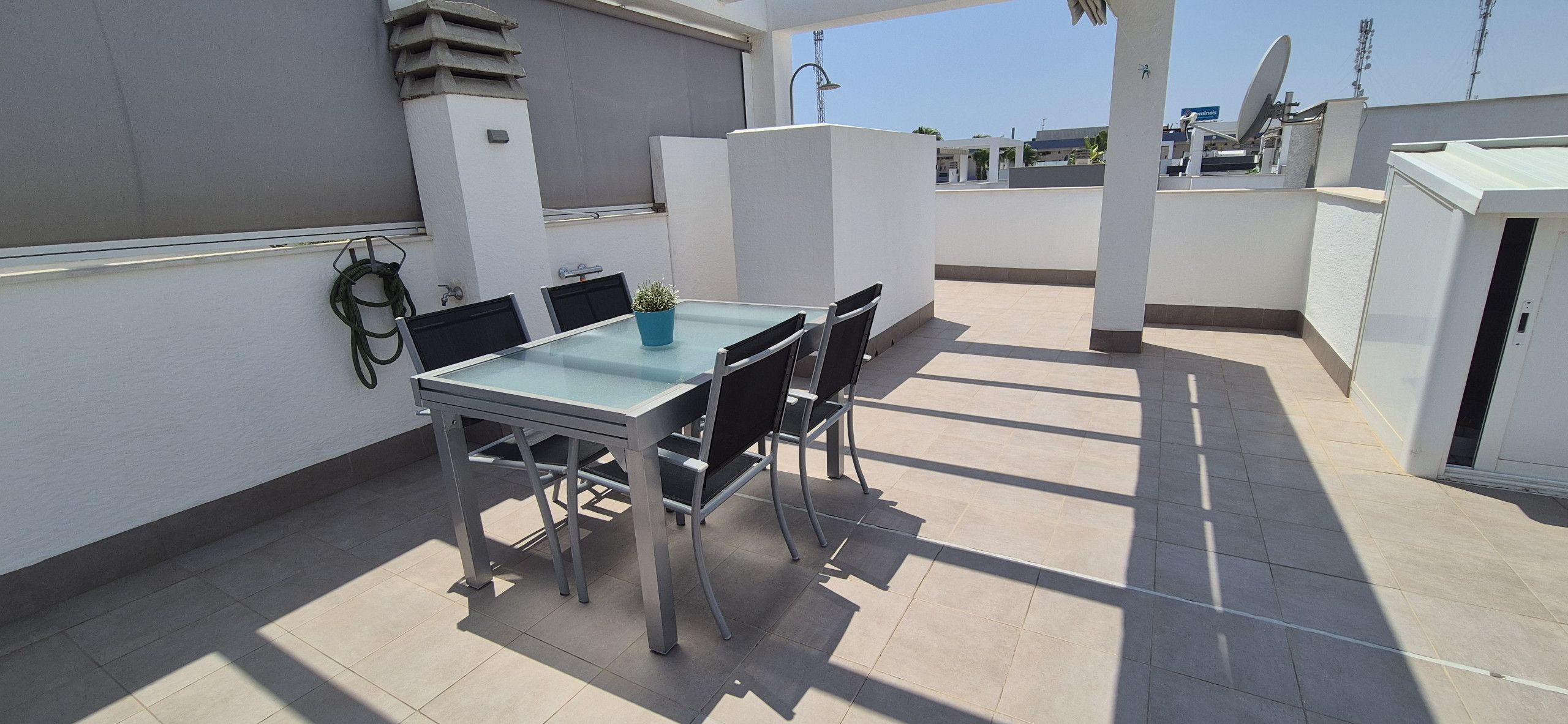 Una terraza en la azotea con una mesa y sillas de vidrio, una planta en maceta y un armario blanco.
