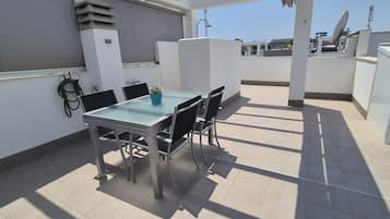 Una terraza en la azotea con una mesa y sillas de vidrio, una planta en maceta y un armario blanco.