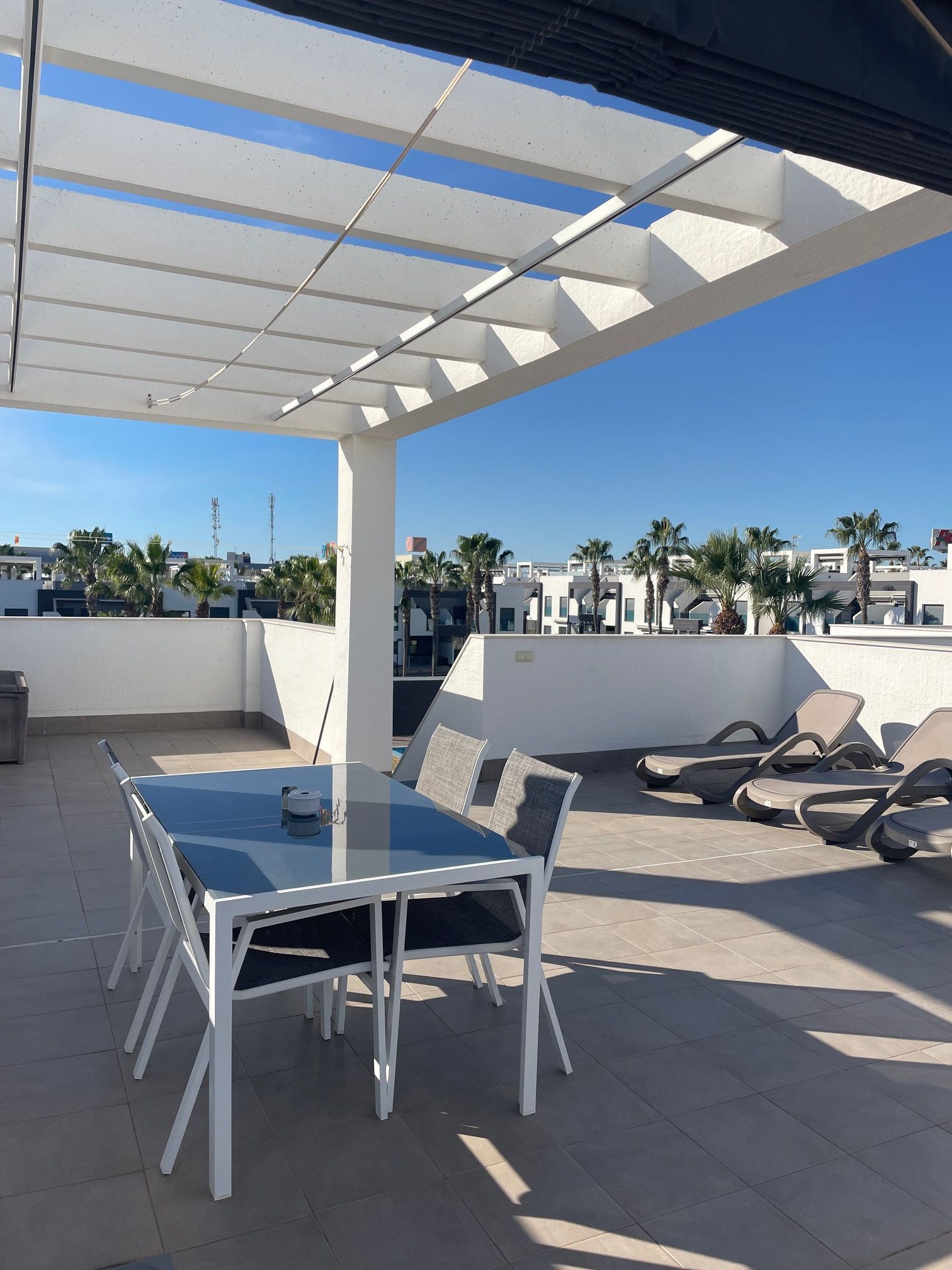 Terraza en la azotea con una mesa y sillas de vidrio, una pérgola y vistas a palmeras y edificios.
