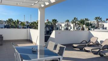 Terraza en la azotea con una mesa y sillas de vidrio, una pérgola y vistas a palmeras y edificios.