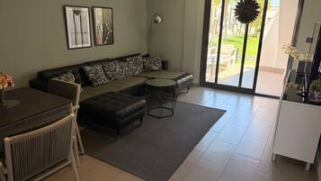 Una sala de estar con un sofá seccional, una mesa de comedor con sillas, un radiador y un ventanal con vistas al exterior.