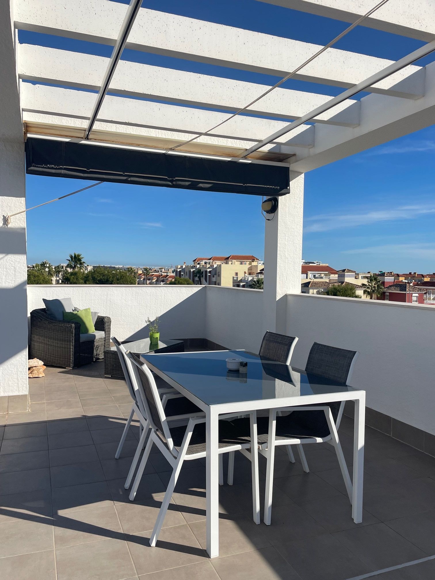 Un patio techado al aire libre con una mesa y sillas de vidrio, un sofá de mimbre y vistas de la ciudad.