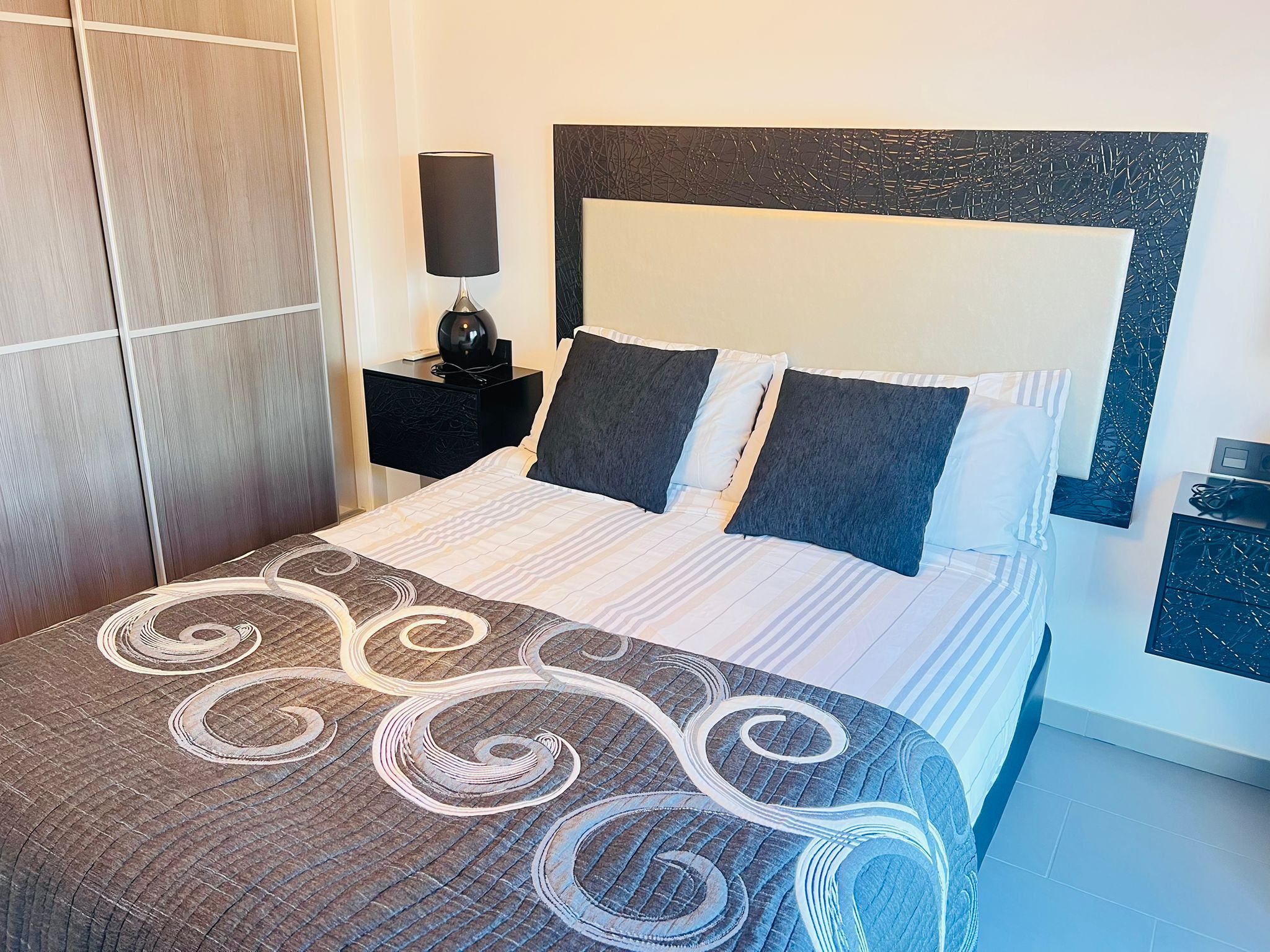 Una cama bien tendida con una colcha estampada, una mesita de noche con una lámpara y una obra de arte colgada en la pared.