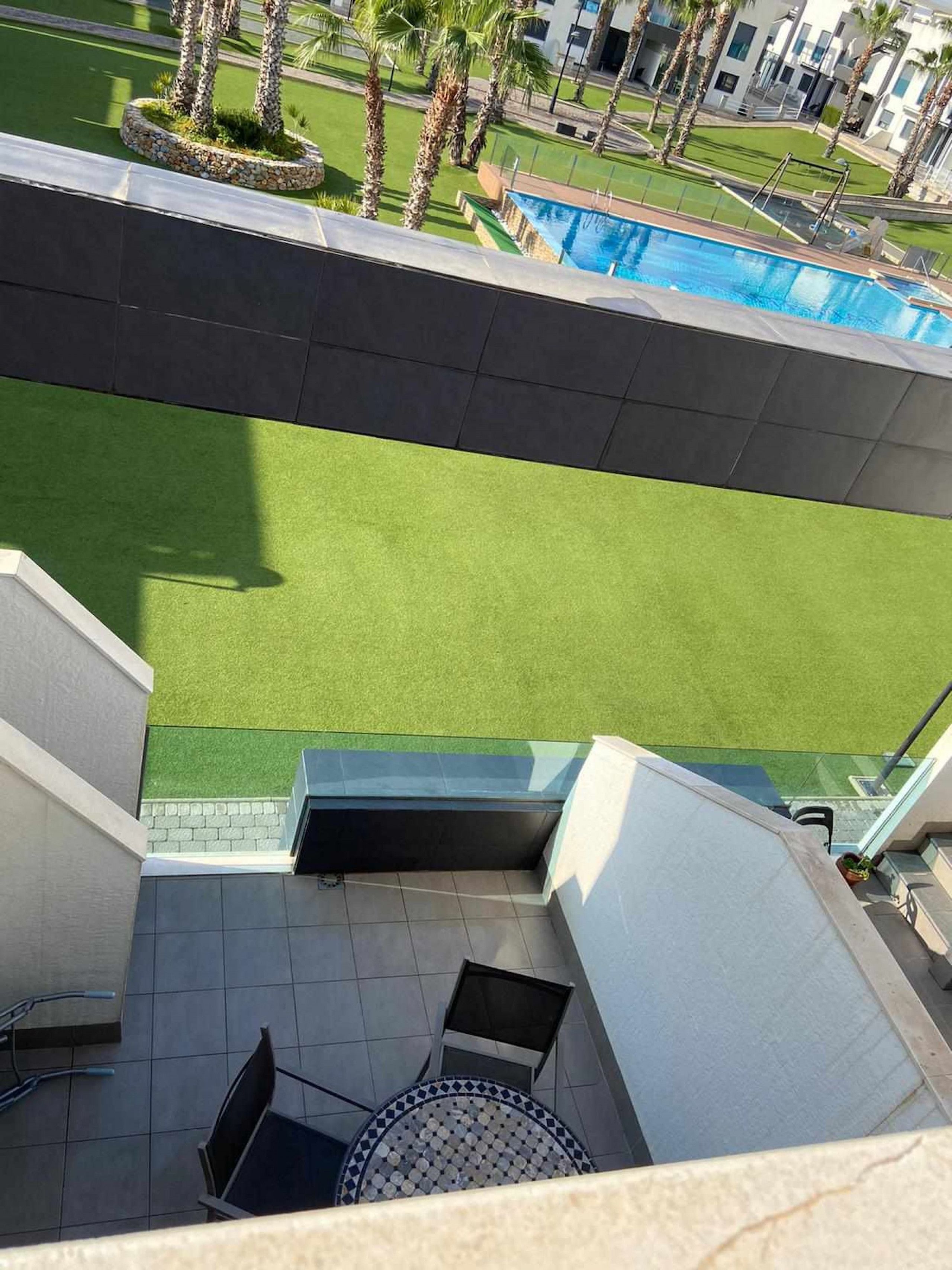 A rooftop terrace with a view of a swimming pool, green lawn, and palm trees.