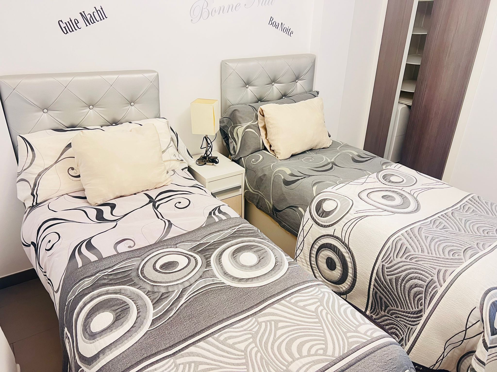 A hotel room with two beds, each with a tufted headboard and patterned bedding. There is a nightstand with a lamp and a small table between the beds.