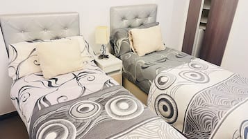 A hotel room with two beds, each with a tufted headboard and patterned bedding. There is a nightstand with a lamp and a small table between the beds.