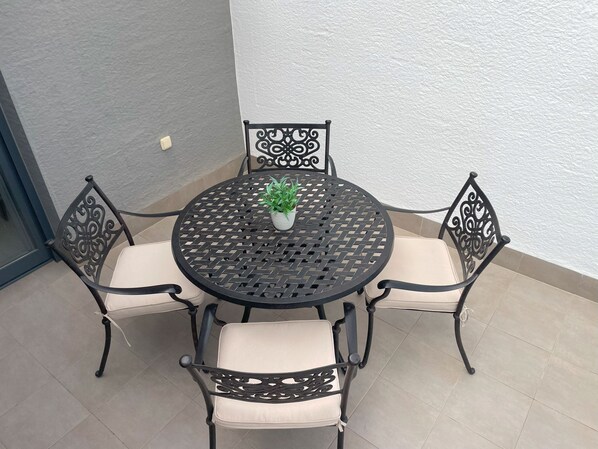 A round metal table with a woven top, surrounded by four cushioned chairs with intricate back designs. A potted plant is placed on the table.