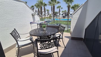 Un balcón con una mesa redonda y sillas, una planta en maceta y vistas a una piscina y palmeras.