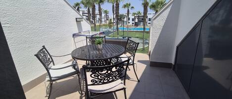 A balcony with a round table and chairs, a potted plant, and a view of a swimming pool and palm trees.