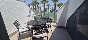 Un balcón con una mesa redonda y sillas, una planta en maceta y vistas a una piscina y palmeras.