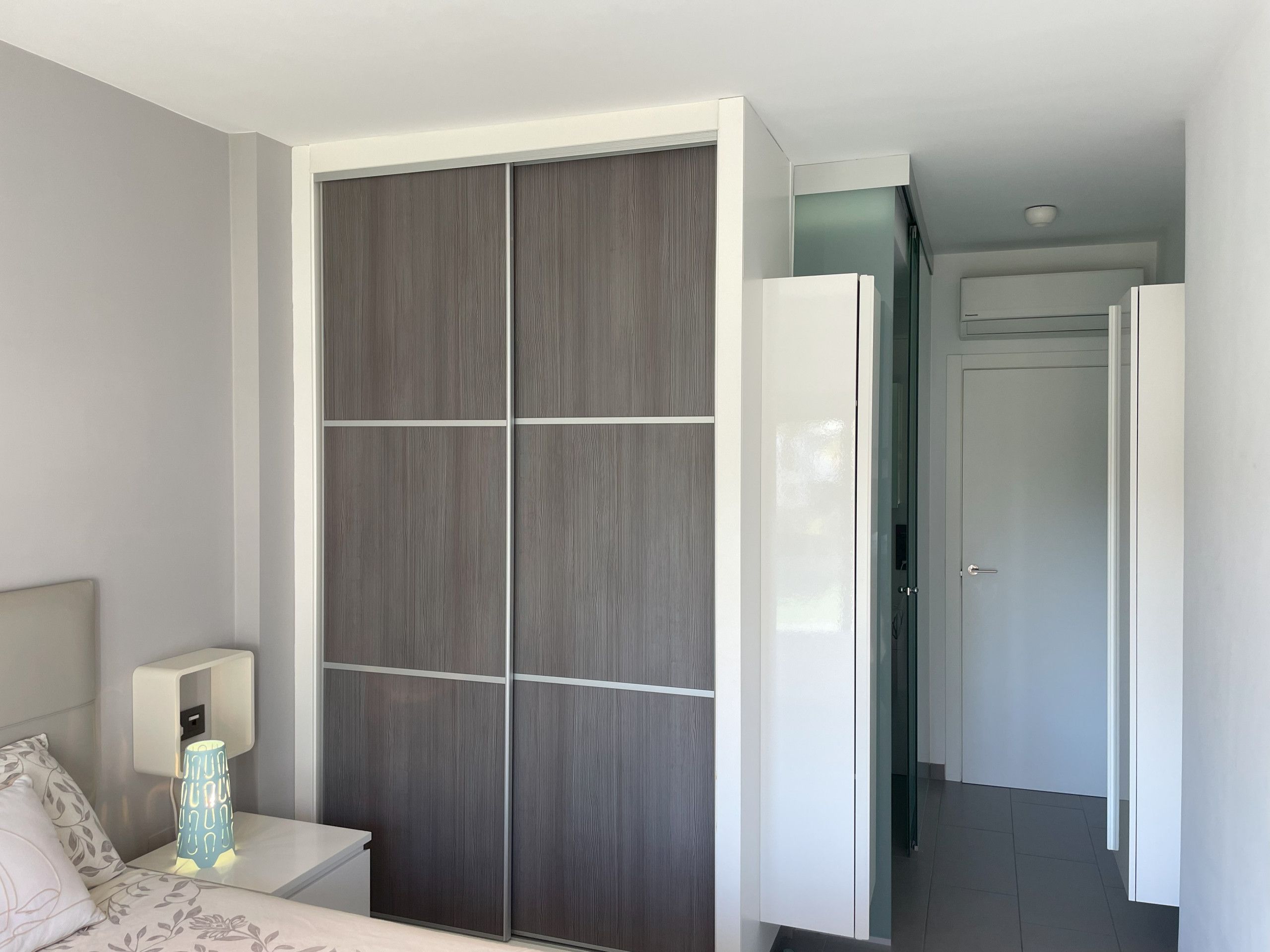 Un dormitorio con cama, mesita de noche, lámpara, armario y una puerta que da a otra habitación.