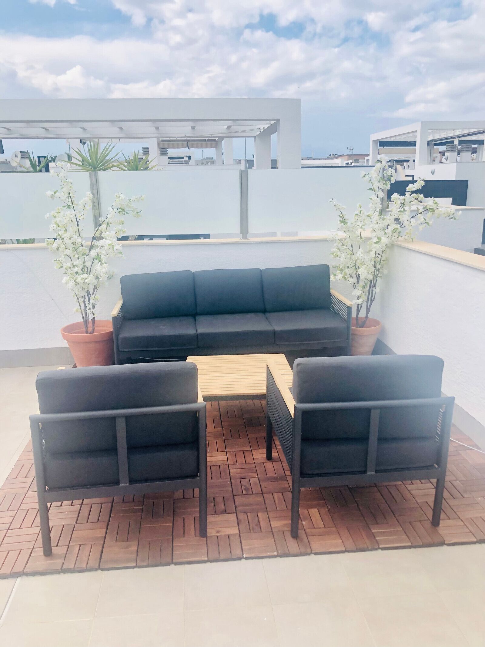 Un área de estar al aire libre con un sofá negro, dos sillones y una mesa de madera. Hay dos plantas en macetas con flores blancas. El mobiliario está sobre una terraza de madera con un diseño en espiga.