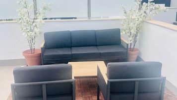 Un área de estar al aire libre con un sofá negro, dos sillones y una mesa de madera. Hay dos plantas en macetas con flores blancas. El mobiliario está sobre una terraza de madera con un diseño en espiga.
