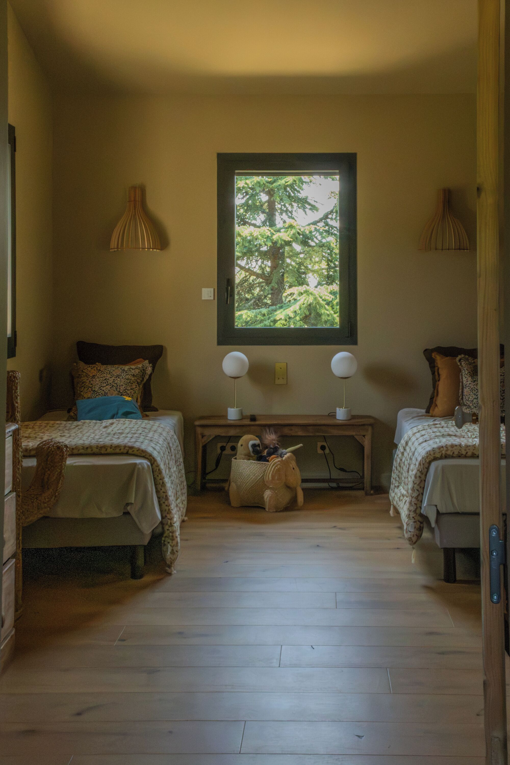 A room with two beds, a window with a view of trees, a desk with lamps, and a small basket with a stuffed toy.