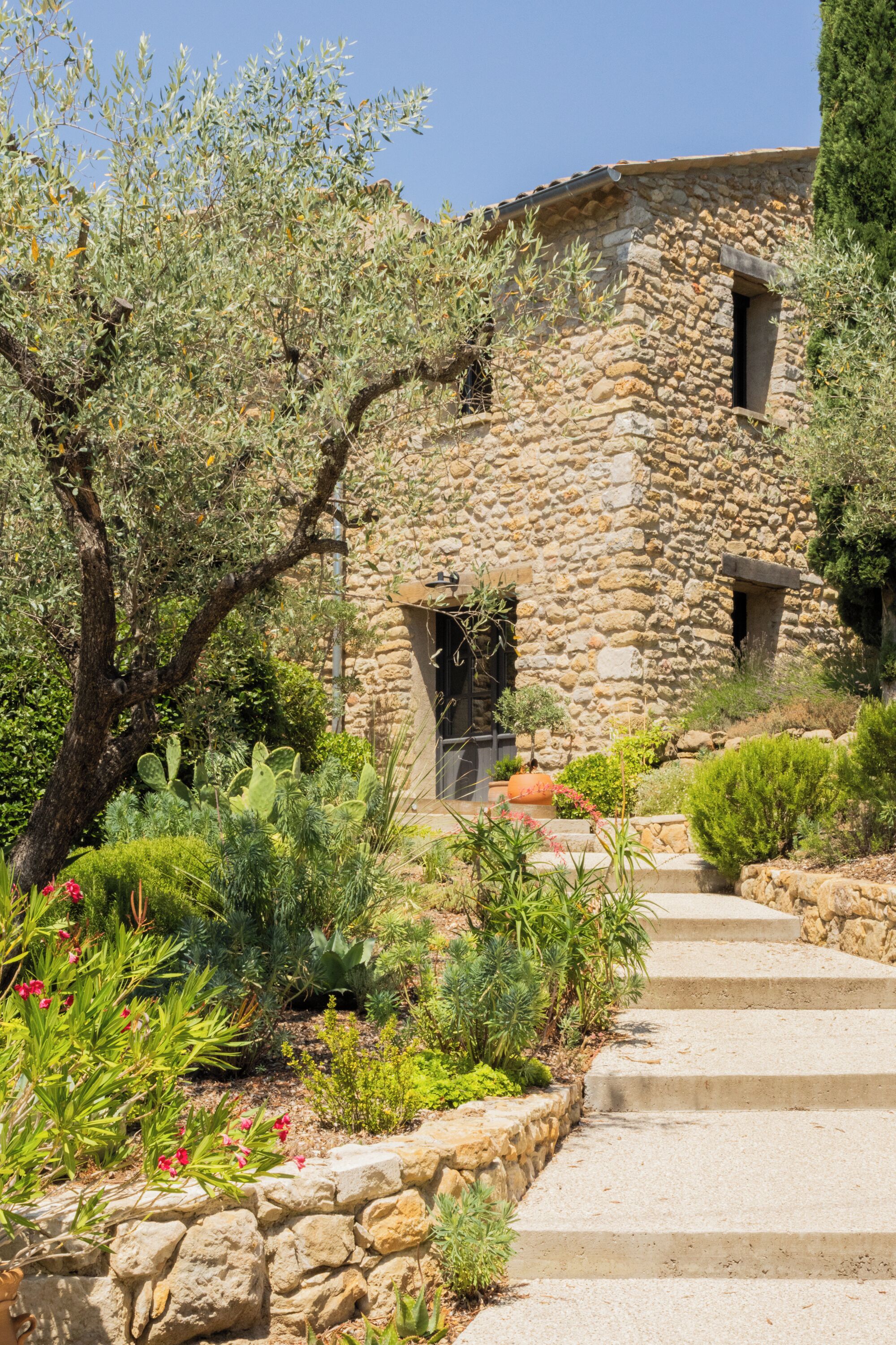 A stone building with a garden in front, featuring a variety of plants and a stone pathway leading to the entrance.