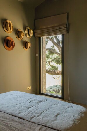 Una habitación con cama, ventana con vista a árboles y varios sombreros en la pared.