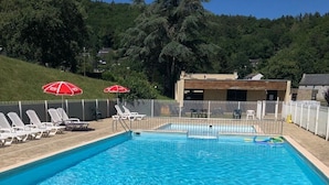 Una piscina rectangular con agua azul cristalina, rodeada por una valla blanca. Se observan sillas de descanso blancas y sombrillas rojas al fondo.