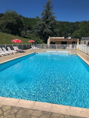 Una piscina rectangular con agua azul cristalina, rodeada por una valla blanca. Se observan sillas de descanso blancas y sombrillas rojas al fondo.