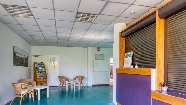 Un área de recepción con mostrador púrpura, sillas de mimbre, una mesita y un tablón de anuncios con varios avisos.