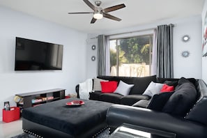 A modern living room with a flat-screen TV, a ceiling fan, a black leather sofa, and a coffee table. - Sleek 5bed pet friendly & pool (Peoria)