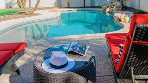 Un área de piscina con una mesa redonda de mimbre, sillas reclinables rojas y una revista.