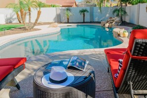 A pool area with a round wicker table, red lounge chairs, and a magazine. - Sleek 5bed pet friendly & pool (Peoria)