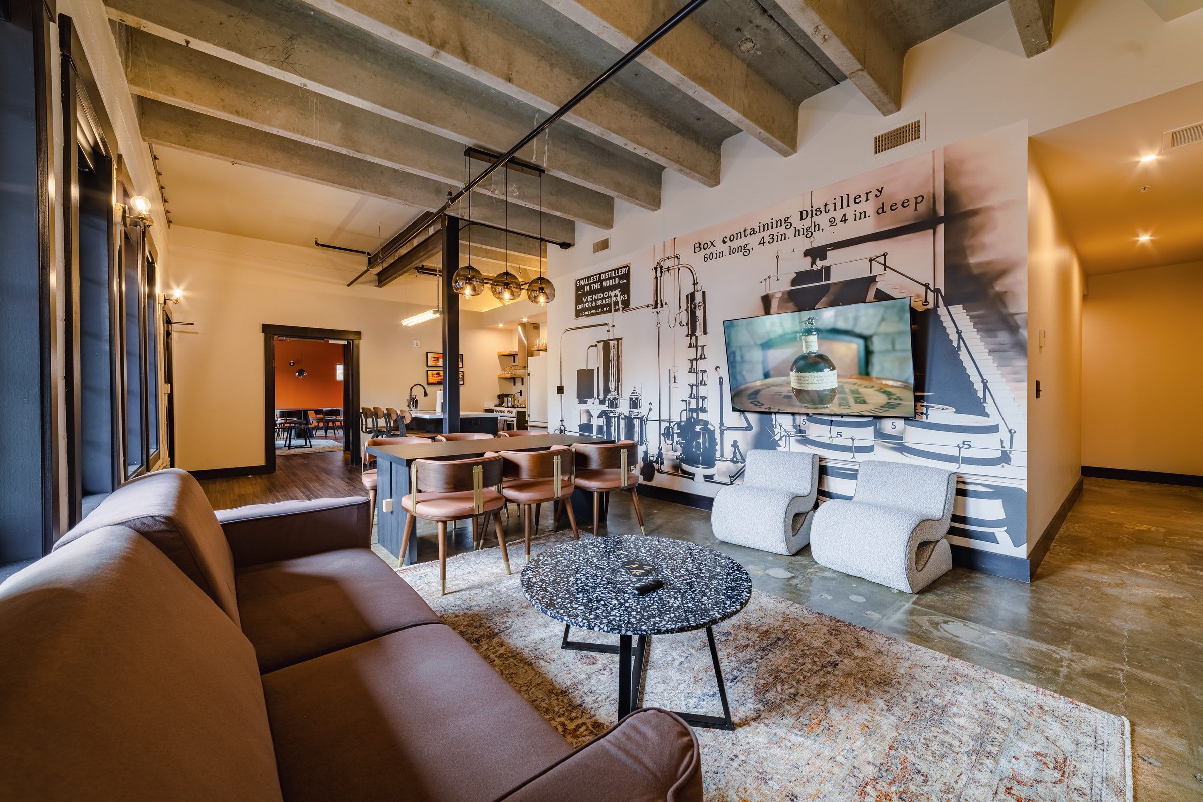 A modern living room with a sofa, a coffee table, and a dining area with chairs. There is a large wall mural depicting distillery equipment.