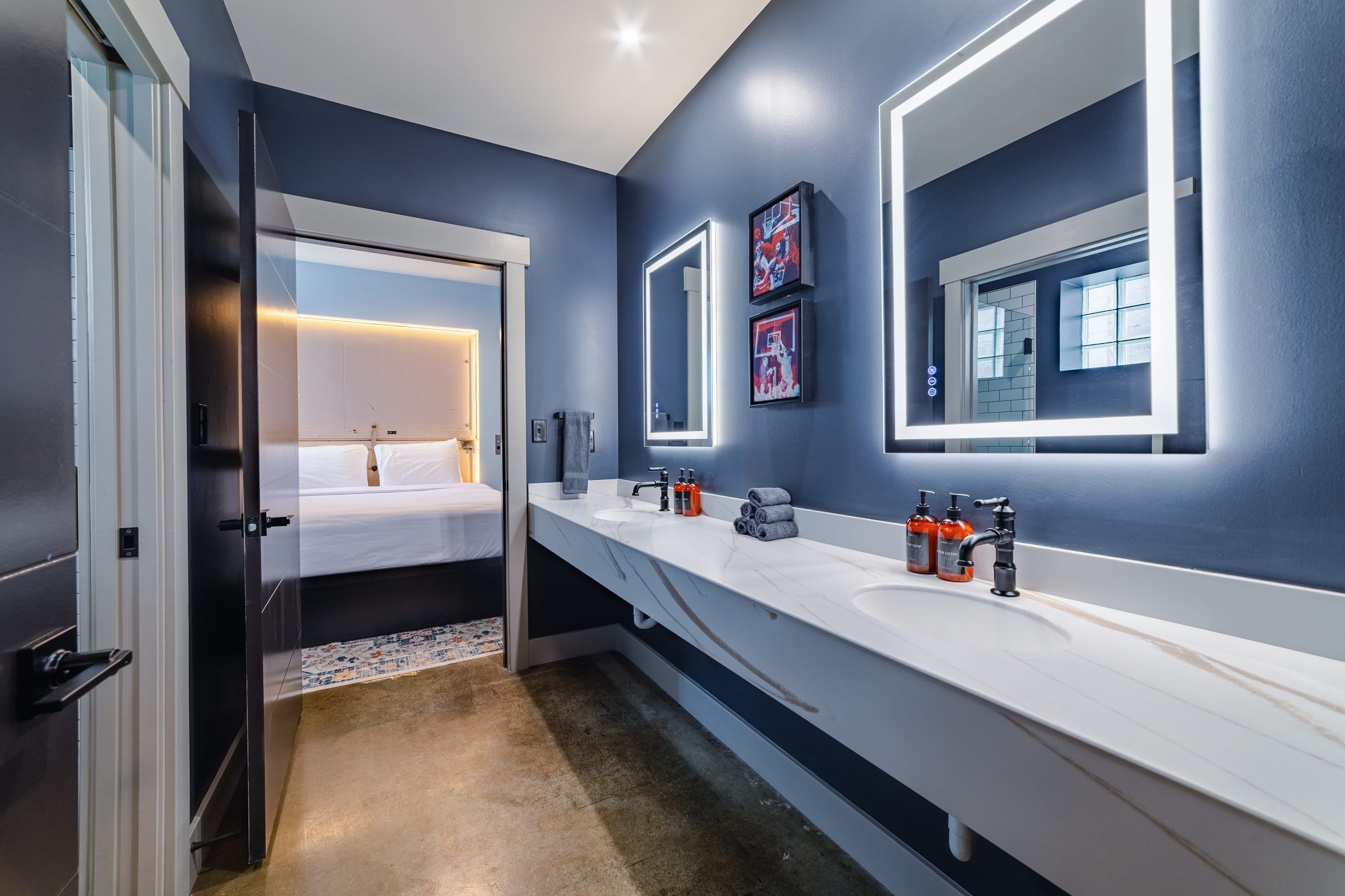Une salle de bain moderne avec un long comptoir blanc, un grand miroir et un lit visible à travers une porte ouverte.